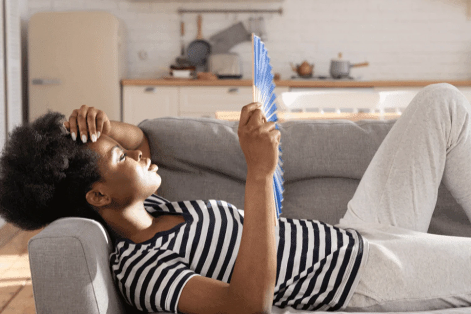 Image of a person feeling hot and cooling off with a blue fan in a well-lit kitchen, illustrating tips on how to stay cool when the AC is broken.