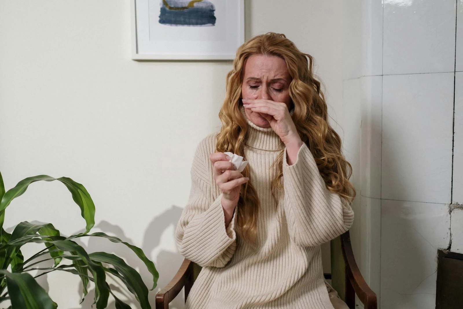 A woman suffering from allergies or a cold in an indoor setting with a potted plant, highlighting the potential need for an air conditioning unit to filter out airborne irritants and improve indoor air quality.